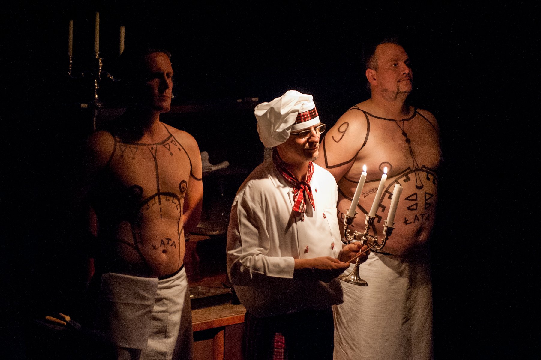 Na zdjęciu trzech mężczyzn: jeden, w środku, ubrany w biały uniform z biała kucharską czapką na głowie, trzyma w ręce świecznik, dwóch pozostałych z nagimi wytatułowanymi torsami za nim.