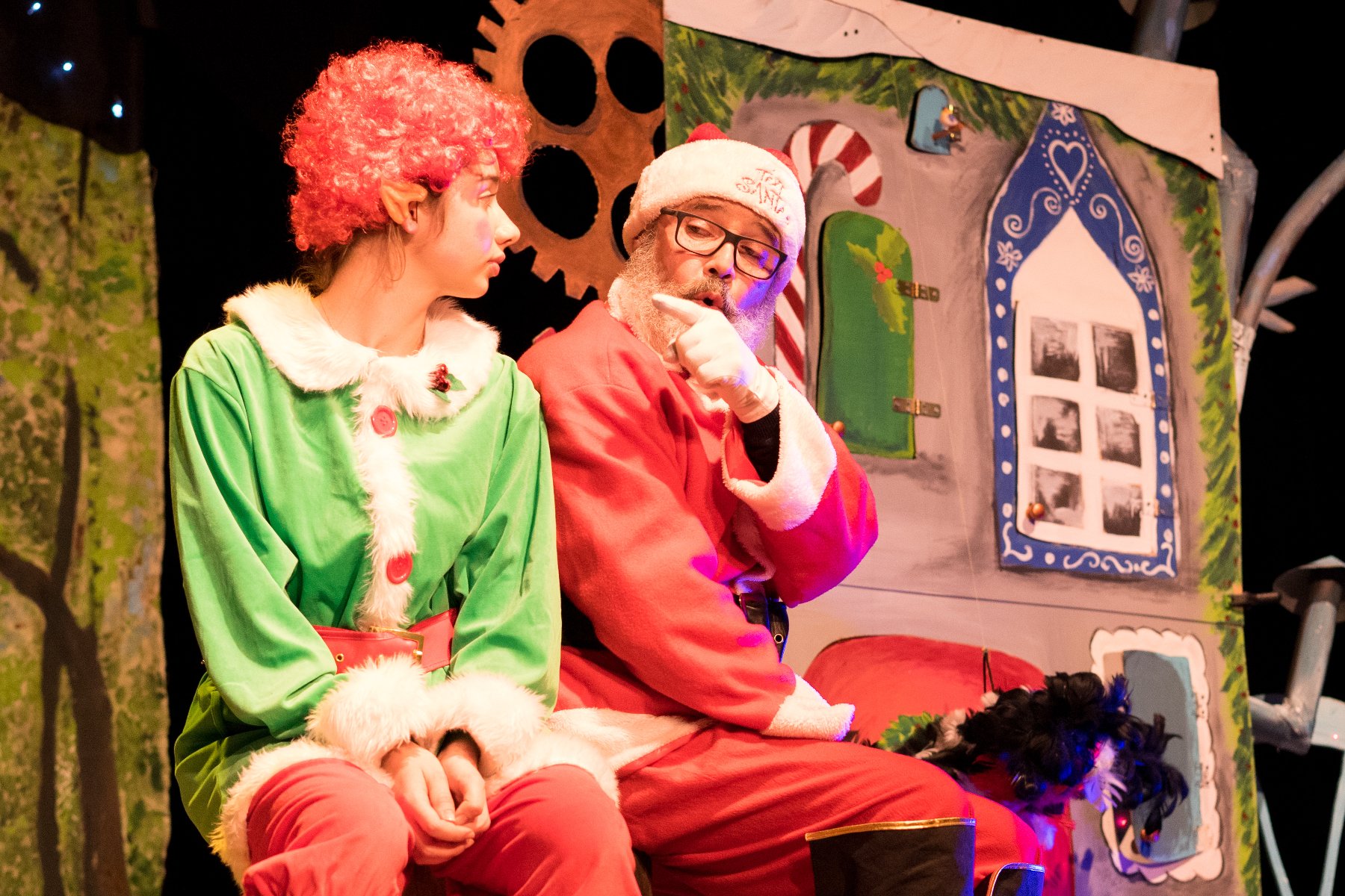 Zdjęcie ze spektaklu: obok siebie siedzą świety Mikołaj i elf, Mikołaj wskazuje palcem na elfa, coś mu tłumaczy. W tle elementy świątecznej scenografii.