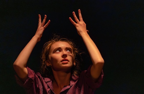 Kobieta w teatralnej pozie z uniesionymi rękoma do góry, ciemne tło.