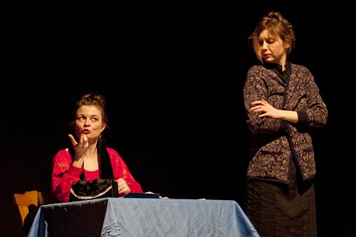 Dwie kobiety, jedna siedzi przy stole i gestykuluje, druga stojąc z założonymi rękoma patrzy w jej kierunku. Ciemne tło.