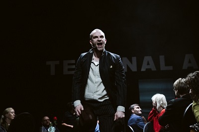 Mężczyzna, który stoi z otwartymi ustami, wokół niego siedzące na krzesłach osoby. Ciemne tło.