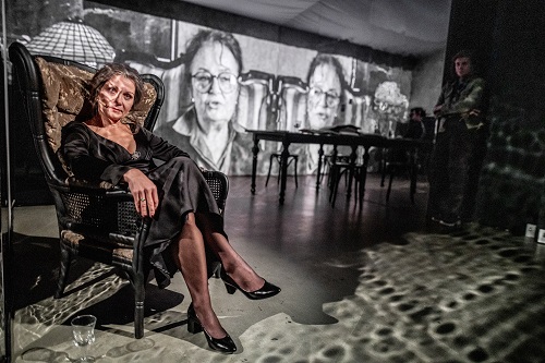 Kobiera w czarnej sukni siedzi na fotelu, za nią czarno-białe fotografie, stół i postać stojąca w cieniu.