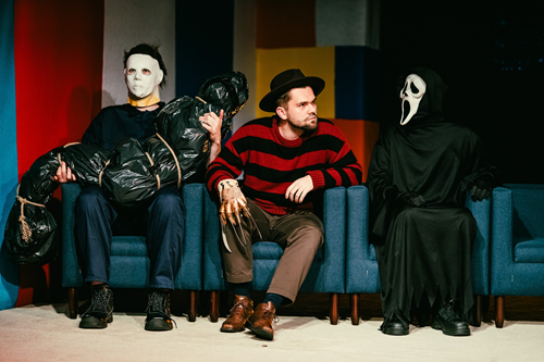 Trzy siedzące na fotelach postacie: w środku mężczyzna patrzący w bok, z jego obu stron postacie w maskach na twarzach.