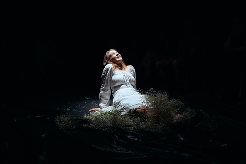 Na czarnym tle zdjęcie młodej, uśmiechniętej dziewczyny w białej sukni. Dziewczyna siedzi oparta rękoma o podłogę, patrzy ku górze.