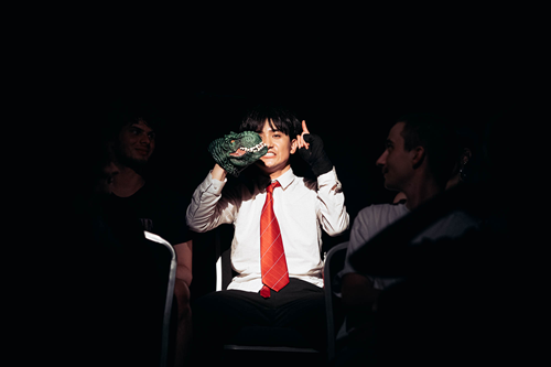 Na ciemnym tle mężczyzna w białej koszuli i czerwonym krawacie siedzi na fotelu, ma uniesione ręce.