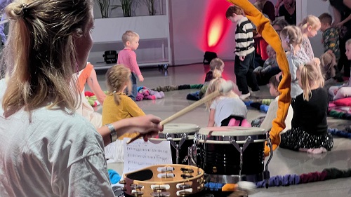 Na pierwszym planie kobieta grająca na perkusji odwrócona tyłem do obiektywu, na drugim planie kilkoro dzieci bawiących się na podłodze.