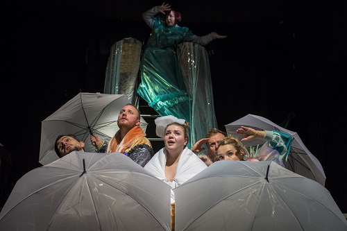 Kilka osób za rozłożonymi parasolami, patrzą w górę. Za nimi kobieta w zielonym połyskującym stroju.