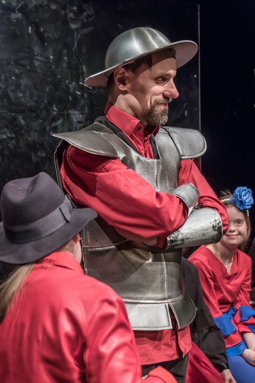 Mężczyzna w blaszanym kapeluszu - hełmie na głowie i stroju przypominającym zbroję, pod którą ma czerwoną koszulę. Ma ręce założone na piersi i delikatnie uśmiecha się.