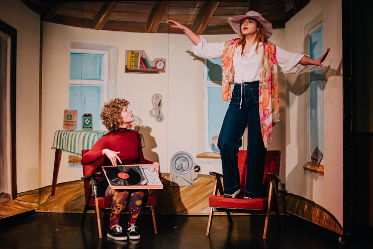 Dwie młode dziewczyny, jedna siedzi i trzyma na kolanach płyę winylową, druga stoi z rozłożonymi rękoma na fotelu.