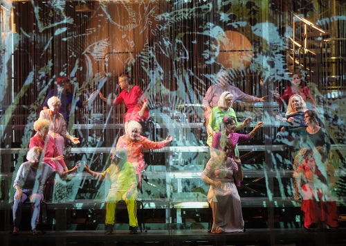 Na zdjęcie kilkanaście osób w kolorowych strojach, siedzą na stopniach w czterech rzędach, zwróceni parami do siebie, niektórzy z wyciągniętymi rękoma.