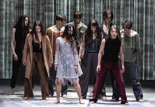 Grupa tancerzy w różnorodnych strojach, wszyscy stoją z opuszczonymi głowami.