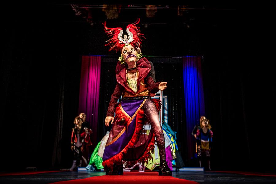 Na zdjęciu postać ubrana w kolorowy strój z maską na twarzy. Stoi na scenie na pierwszym planie, za nią w tle inne postacie.