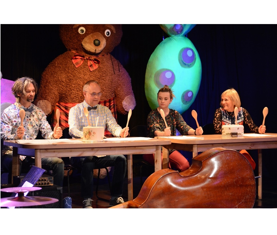 kadr ze spektaklu przedstawia czworo aktorów siedzących przy stołach i trzymających uniesione drewniane łyżki. Przed nimi leżą odwrócone do góry dnem garnki.