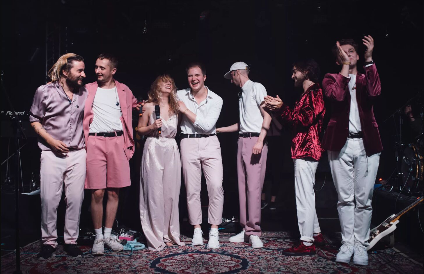 Siedmioro ludzi ubrani w odcienie różowego stoją na scenie wyłozonej dywanami. Za nimi instrumenty muzyczne.
