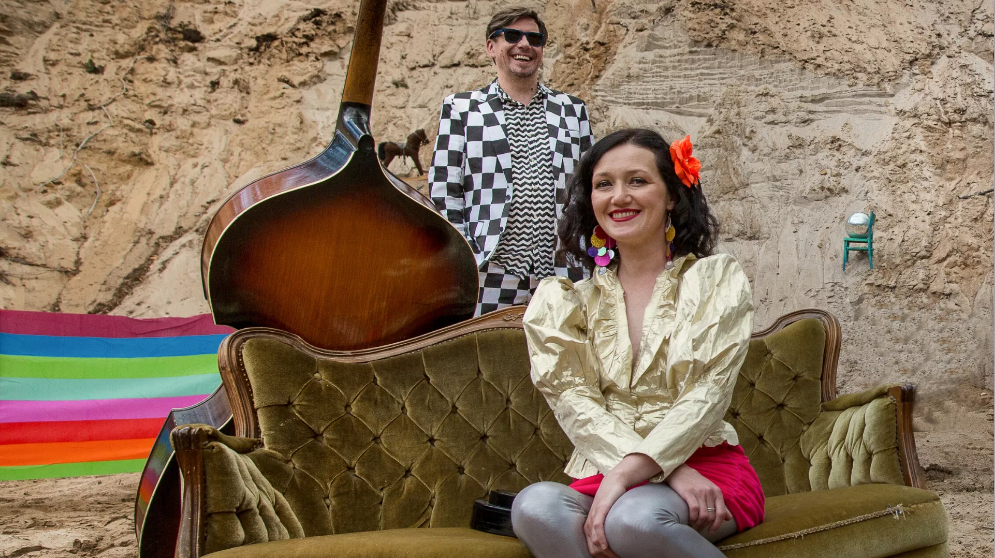 Kobieta w złotej marynarce siedzi na zielonej kanapie na plaży. Za nią mężczyna ubrany w garnitur w szachownicę. O kanapę oparty jest kontrabas. W tle kolorowy parawan w paski i ściana skalna.