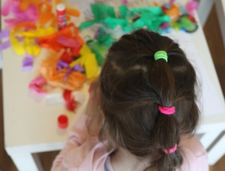 Zdjęcie zrobione z góry. Dziewczynka stoi tyłem do obiektywu. Przed nią na stoliku leżą kolorowe przybory plastyczne.