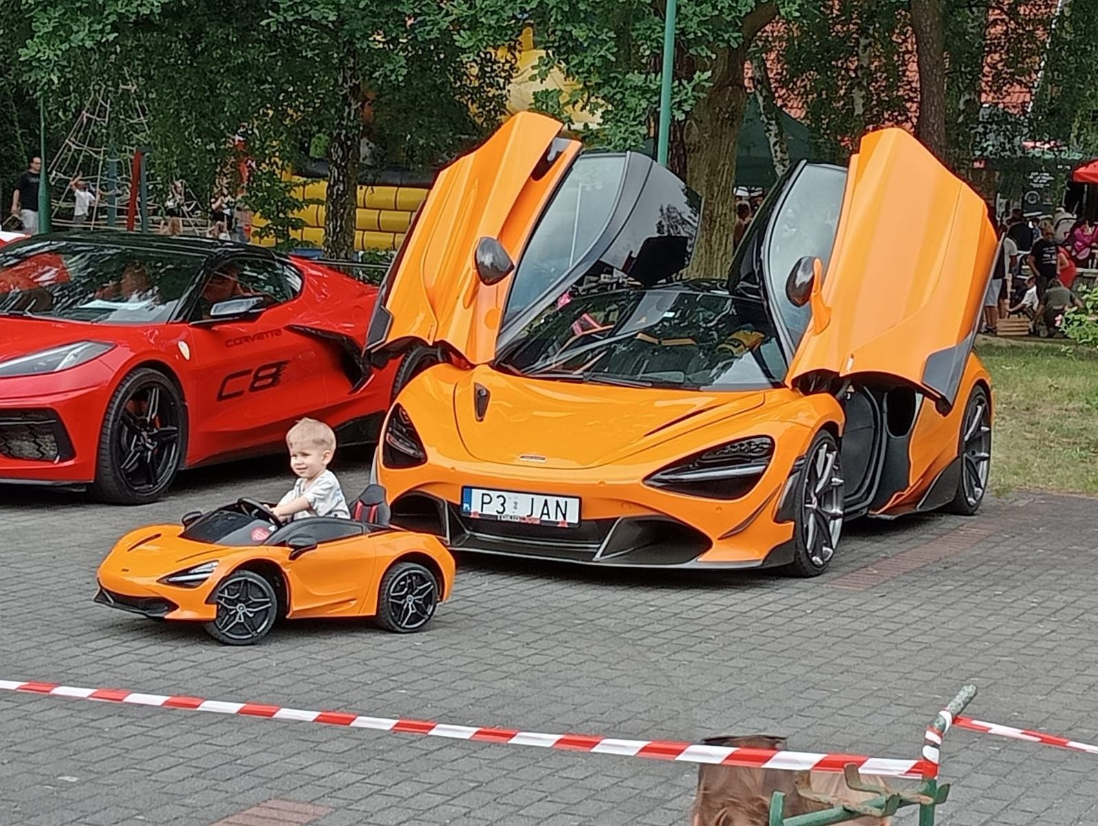 Dwa duże auta i jedno mniejsze, w którym siedzi mały chłopiec. W tle plac zabaw i drzewa.