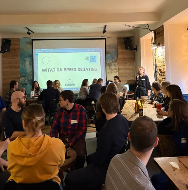 Grupa ludzi siedzi przy stołach w barze, uczestnicząc w wydarzeniu "speed debating". Na ścianie widać duży ekran z napisem "Witaj na speed debating'. Jedna osoba stoi z mikrofonem i przemawia do uczestników, a reszta rozmawia w małych grupach.