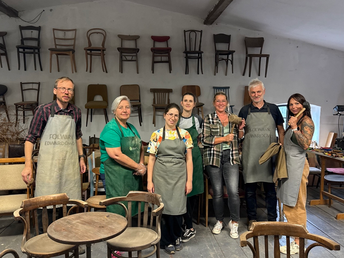 Grupa osób w fartuchach. Wokół przedmioty wykonane z drewna. W tle krzesła wiszące na szarej ścianie.