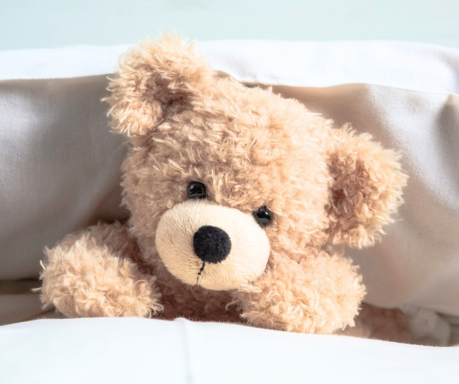 Pluszowy, jasnobeżowy miś leży pod kołdrą, oparty o białą poduszkę