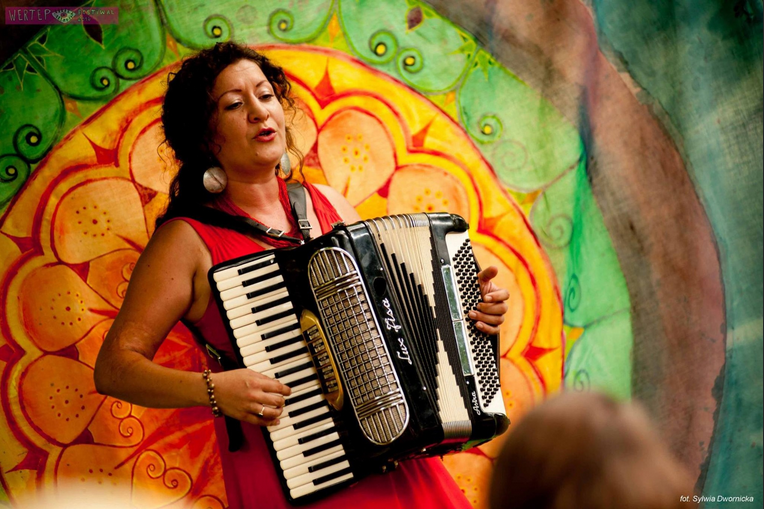Kobieta o ciemnych kręconych włosach, ubrana w czerwoną sukienkę, gra na akordeonie i śpiewa. W tle widoczny jest kolorowy, dekoracyjny wzór w ciepłych odcieniach zieleni, żółci i pomarańczy.