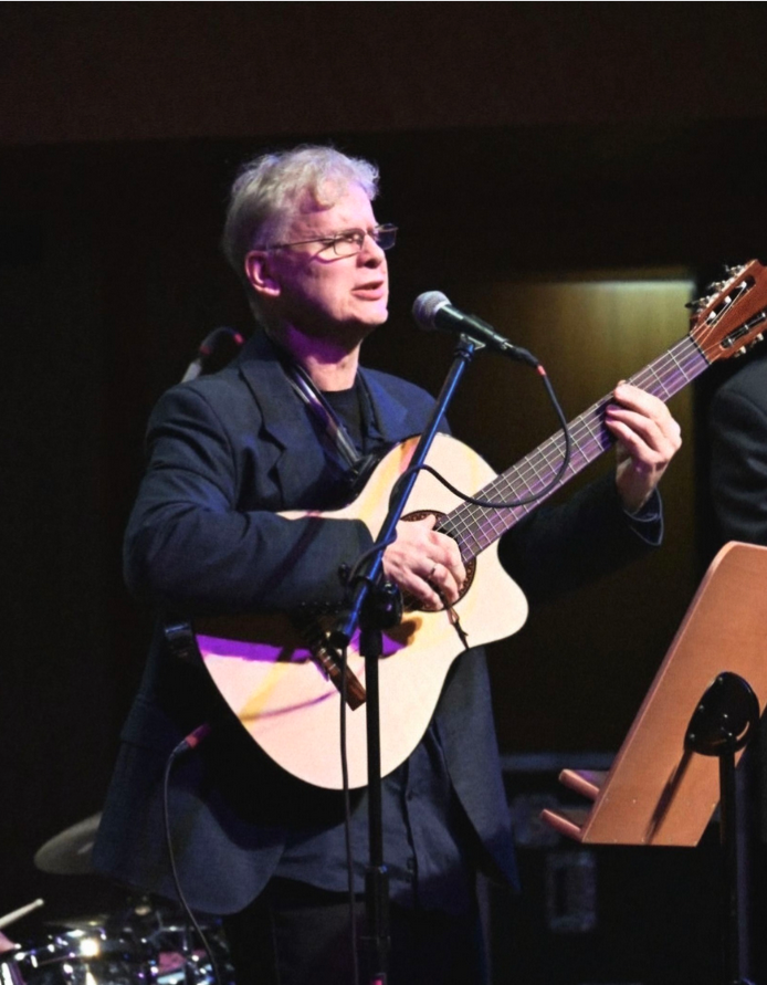 Zdjęcie mężczyzny na scenie, na której gra na gitarze i śpiewa. Przed nim stoi mikrofon na statywie.