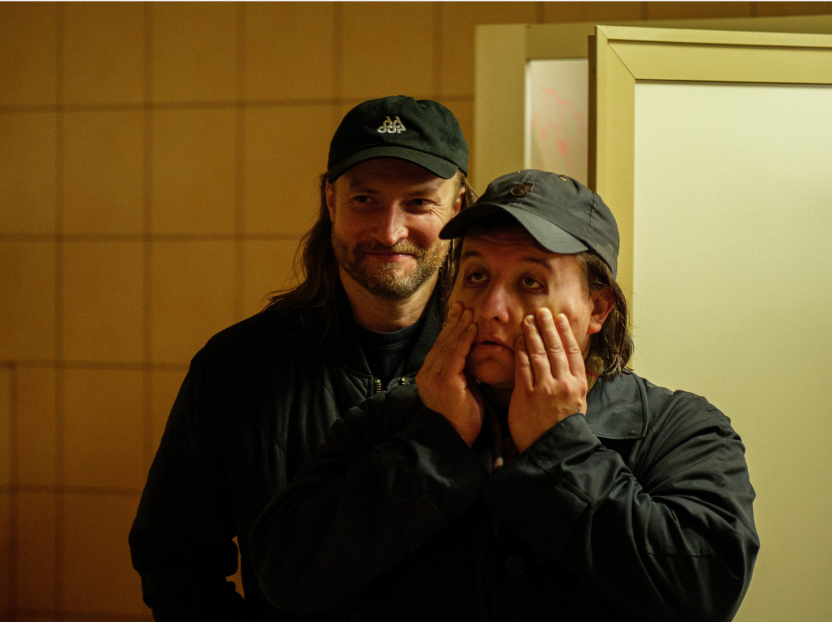 Dwóch mężczyzn w ciemnych ubraniach i czapkach z daszkiem stoi w słabo oświetlonym pomieszczeniu. Mężczyzna z przodu trzyma dłonie na policzkach, wyglądając na zmęczonego lub zrezygnowanego, podczas gdy drugi, stojący za nim się uśmiecha..