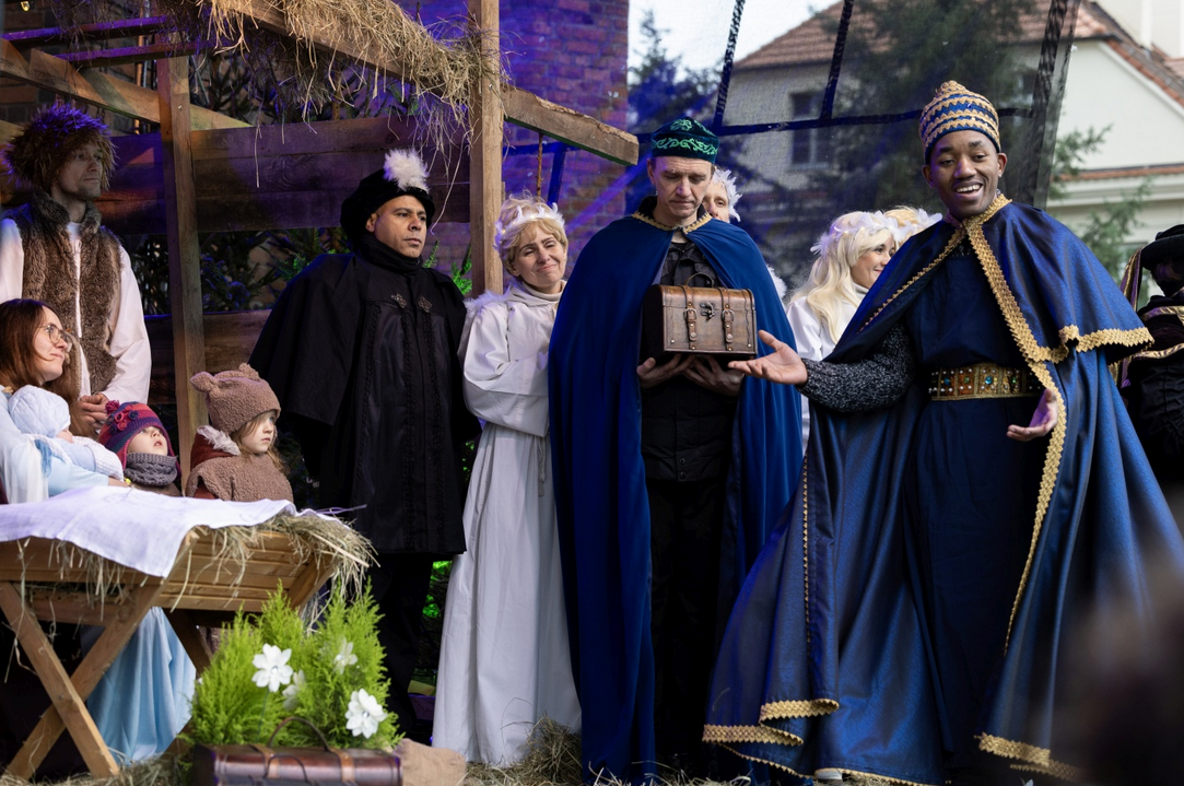 Plenerowa scena jasełek. Widać postacie w historycznych strojach stoją przy drewnianej stajence z sianem. Maria i Józef z dzieckiem w żłóbku, aniołowie oraz trzej królowie, jeden trzyma ozdobną szkatułkę i gestem zwraca się do publiczności.