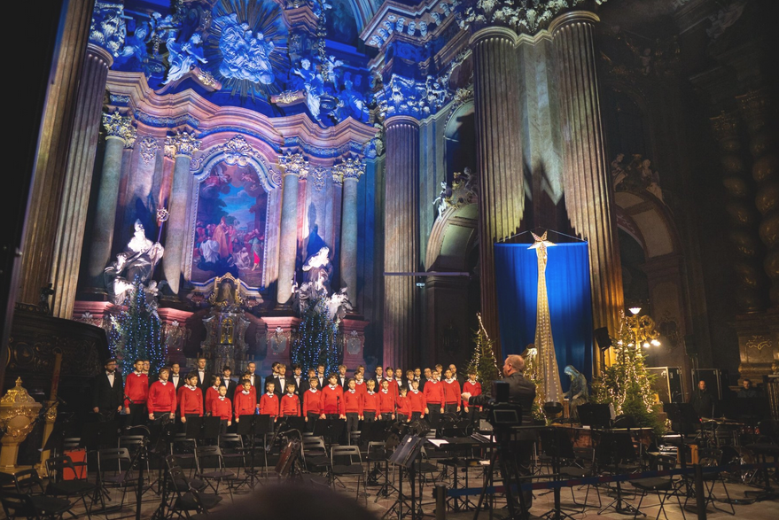 Na zdjęciu chór śpiewający w poznańskiej farze, widać chórzystów, podświetlony ołtarz i dyrygenta.