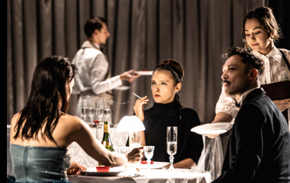 Grupa osób siedzących przy stole, obsługiwanych przez kelnerkę, w tle widac kelnera. Kobieta na środku zdjęcia trzyma w ręce papierosa. Na stole widać kieliszki i butelkę.