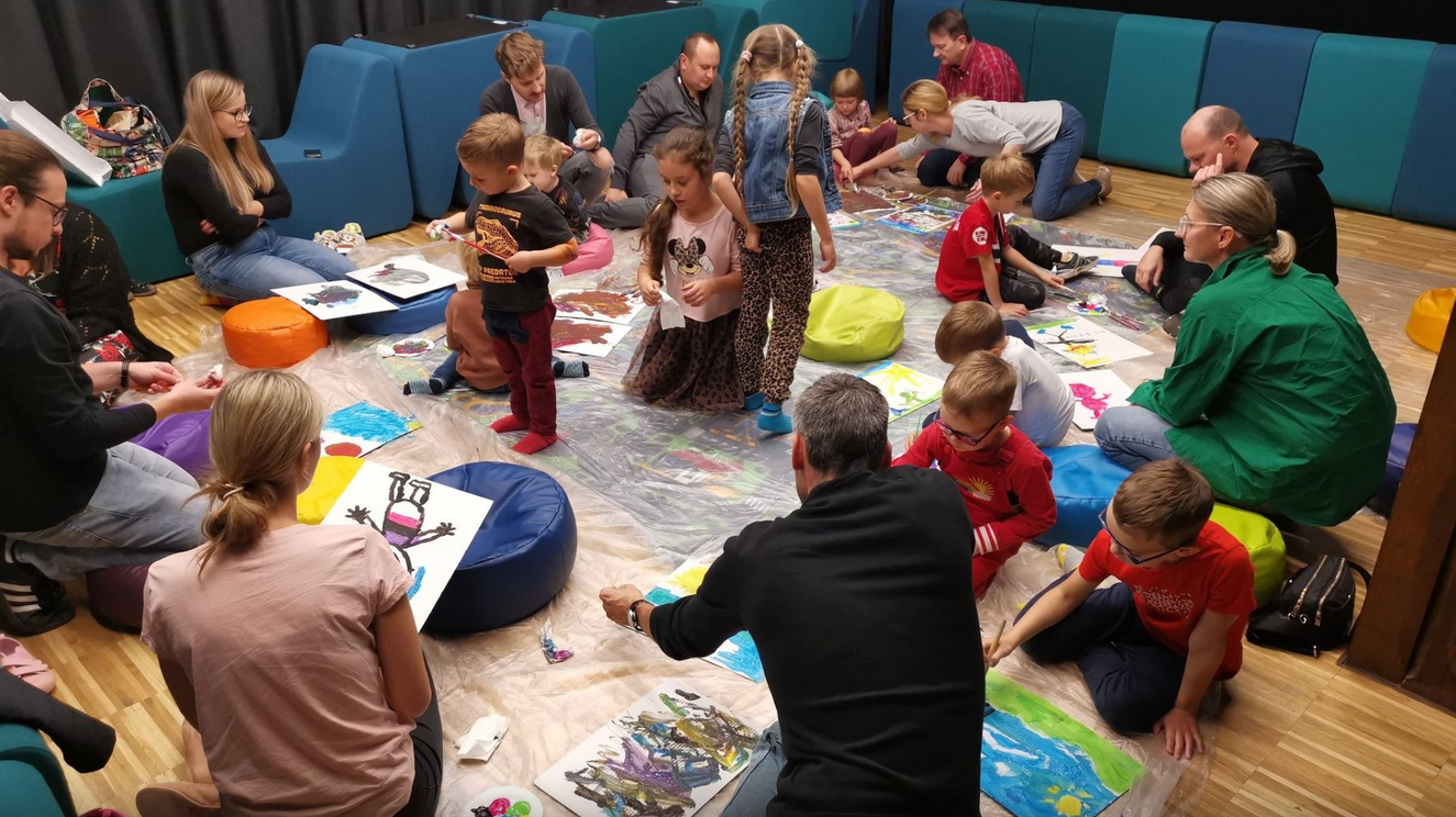 Grupa dzieci i dorosłych siedzi na podłodze w sali warsztatowej, wspólnie malując kolorowe obrazy na kartkach rozłożonych na folii ochronnej. Wokół widać pufy, farby i rysunki w trakcie tworzenia.