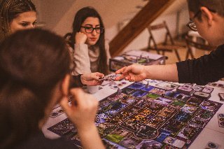 Grupa osób skupiona wokół stołu podczas rozgrywki w rozbudowaną grę planszową. Na pierwszym planie widoczne plecy i profil ciemnowłosej kobiety oraz dłoń gracza sięgająca po kartę na kolorowej, szczegółowej planszy. W centralnej części dwie młode kobiety z uwagą obserwują przebieg gry. Jedna z nich, w okularach i beżowym swetrze opiera podbródek na dłoni. Tło tworzy przytulne, domowe wnętrze z drewnianymi schodami i krzesłami w ciepłym, przygaszonym świetle.