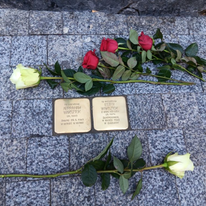 Złote kamienie z imionami i nazwiskami wmurowane w chodnik. Obok nich leżą białe i czerwone róże.