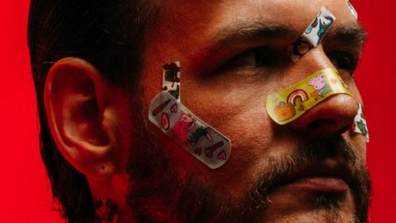A photo of a man's face with colorful plasters glued to his nose, cheeks and forehead. Red background.