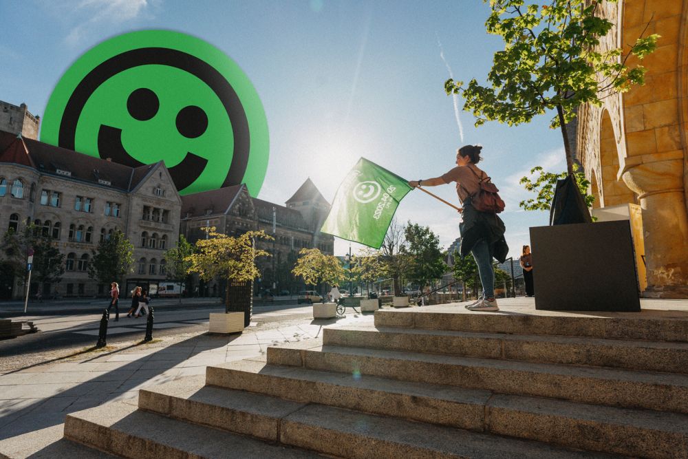 A woman with a green flag in her hands stands on the steps in front of the building. In the background there is a street and a historic building with a smiling face emoticon sticking out from behind it. - grafika artykułu