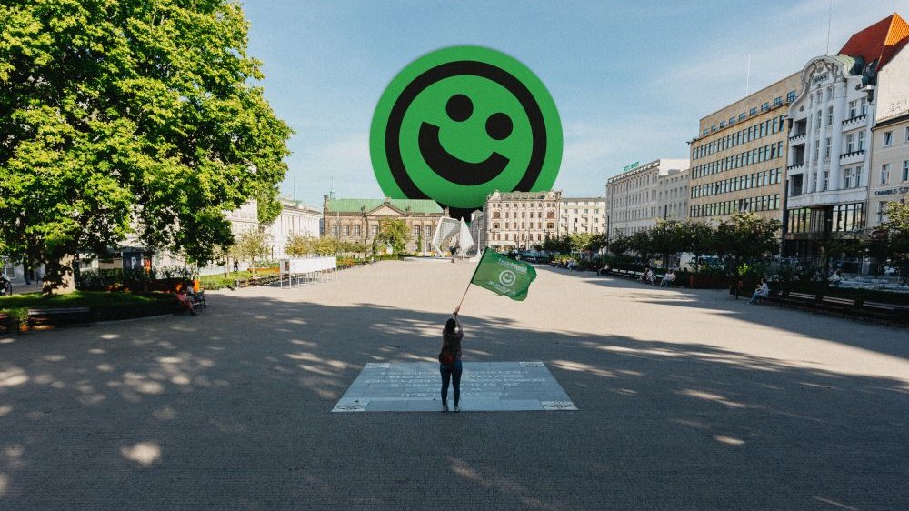 A person standing in a large square waves a green flag. In the distance, there are buildings with a smiling face emoticon sticking out from behind them.