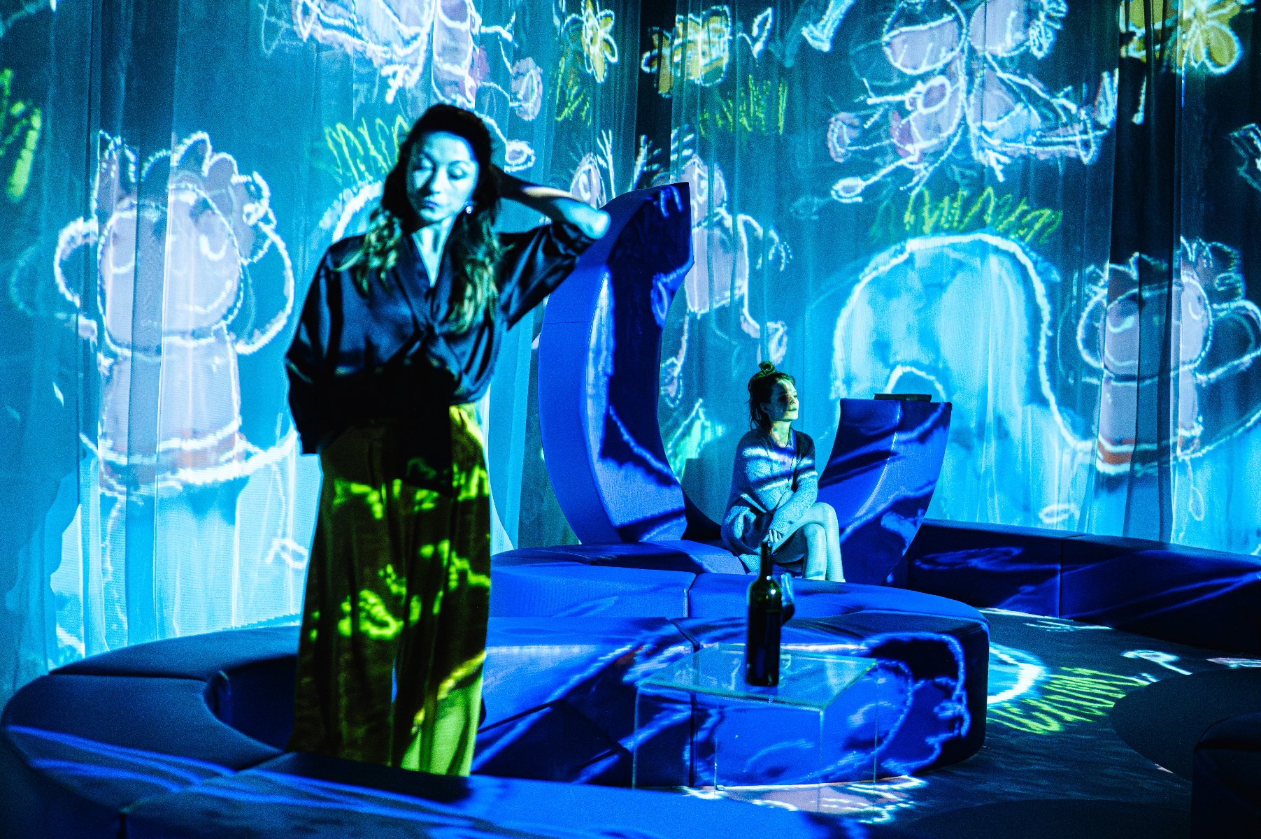 fot. Dawid Stube Dwie młode kobiety w strojach domowych na tle wideo z rysunkami dziecią narysowanych kredą. na stoliku butelka wina.