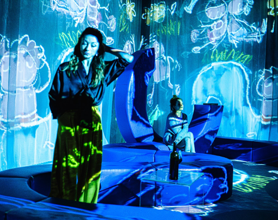 fot. Dawid Stube Dwie młode kobiety w strojach domowych na tle wideo z rysunkami dziecią narysowanych kredą. na stoliku butelka wina.