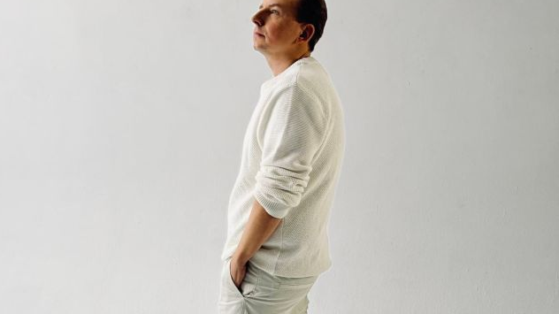 Artysta ubrany na biało (sweter, spodnie i trampki) stoi w wyluzowanej pozie, trzyma ręce w kieszeniach, za nim widać jasną, prawie białą ścianę.