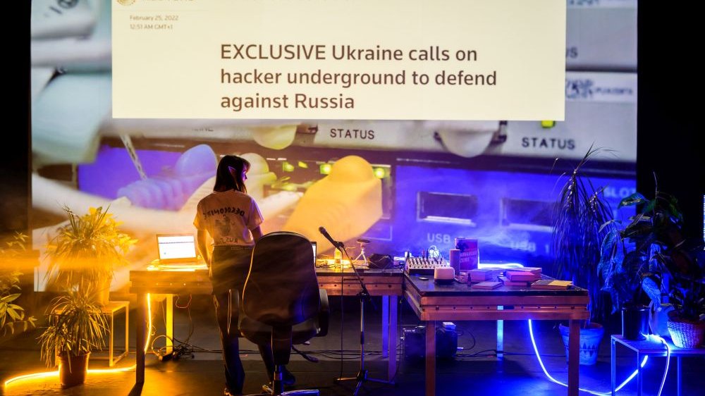 Dziewczyna stoi przed wielkim ekranem z napisem "Exclusive Ukraine calls on hacker undergroung to defend against Russia"