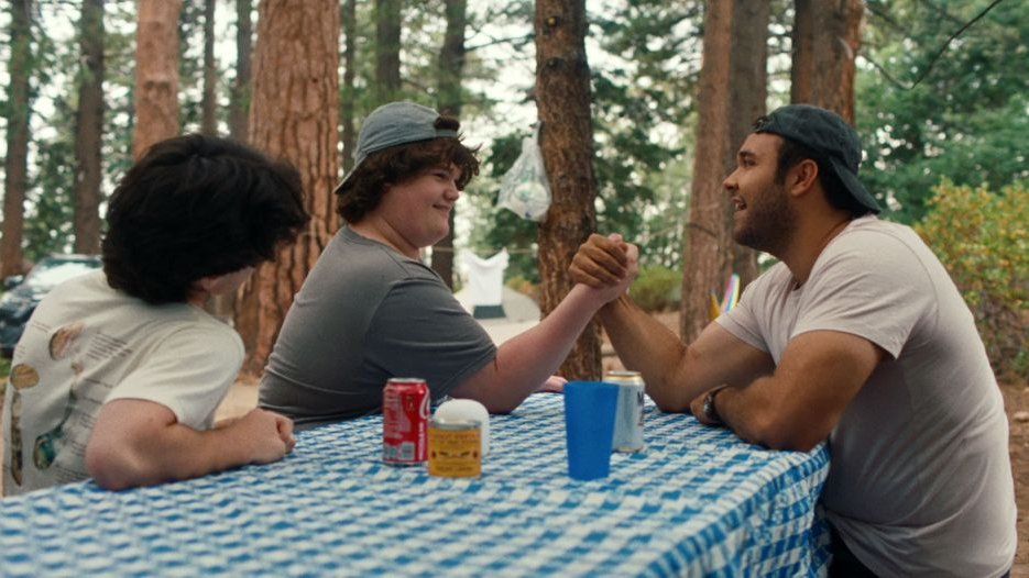 Mężczyzna w czapce z daszkiem siedzi przy stole piknikowym z dwoma chłopcami, z jednym z nich siłuje się na rękę. Dookoła las.