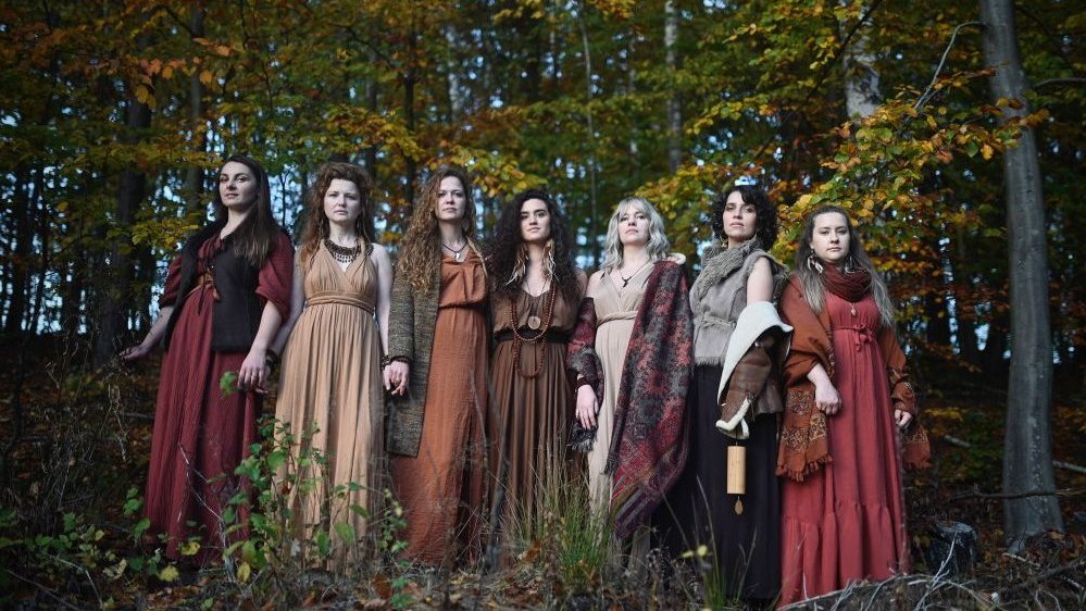 Siedem kobiet w długich włosach i sukniach stoi w lesie.