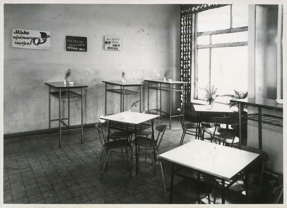 Czarno-białe zdjęcie pomieszczenia z kwadratowymi stolikami i krzesłami. Na ścianie blaszane tabliczki z komunikatami. - grafika artykułu