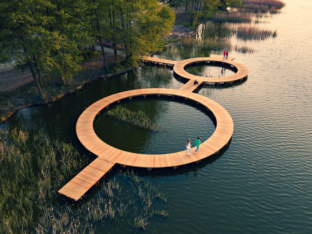 Wizualizacja drewnianego pomostu na wodzie. Pomost ma kształt dwóch połączonych okręgów, chodzą po nim ludzie. - grafika artykułu