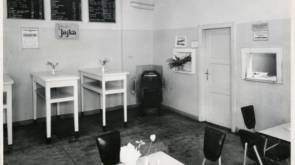 Sala z białymi stolikami i czarnymi krzesłami. W ścianie otwarte okienko do wydawania potraw.