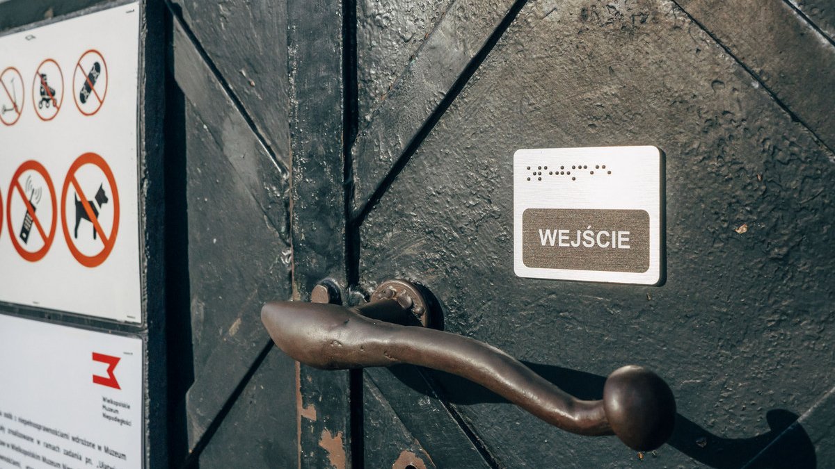 Metalowe drzwi i klamka, na nich niewielka tabliczka z napisem wejście i z wypukłymi kropkami