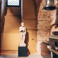 A statue of a knight, next to it there was a pedestal on which a sculpture stood. On the right, a medium-sized bell hangs on the brick wall.