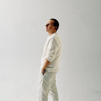 Artysta ubrany na biało (sweter, spodnie i trampki) stoi w wyluzowanej pozie, trzyma ręce w kieszeniach, za nim widać jasną, prawie białą ścianę.
