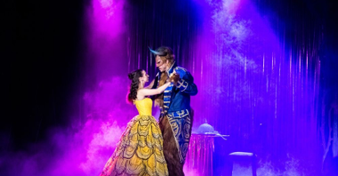 Scena ze spektaklu "Piękna i Bestia" - kobieta w żółtej sukni i mężczyzna w niebieskim stroju, z maską na twarzy i rogami na głowie, tańczą na scenie.