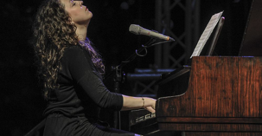 Zdjęcie kobiety grającej na fortepianie. Kobieta ma zamknięte oczy i twarz skierowaną ku górze.